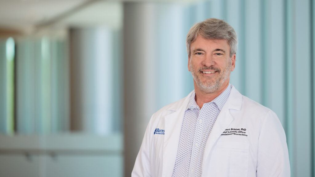 Dr. Robert Bowser is pictured in a brightly lit lobby of the Barrow Neuroplex. He is wearing a white lab coat and smiling at the camera.